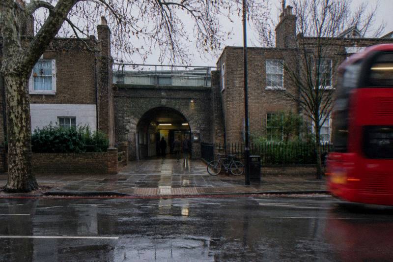 Rainy Mile End street in east London, puddles by the station entrance, red double-decker passing