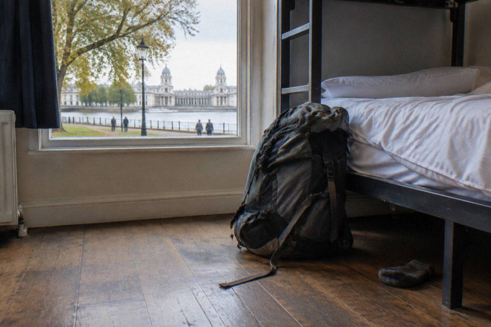 A backpacker rucksack sitting on a wooden floor beside a lower bunk bed with fresh white bedding