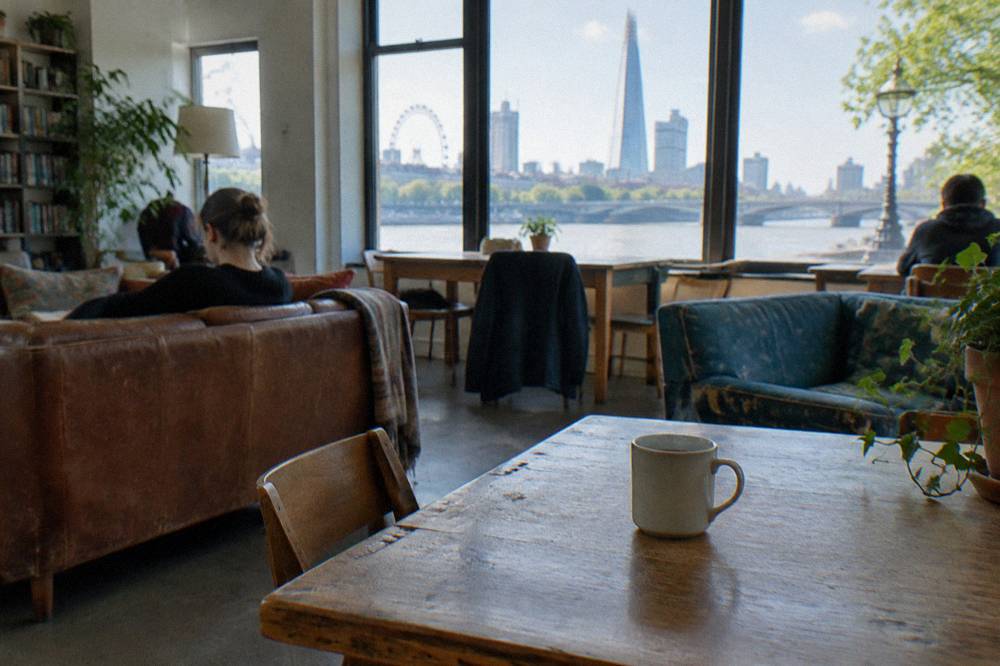 A bright hostel common room with wooden tables, sofas and large windows onto a London street