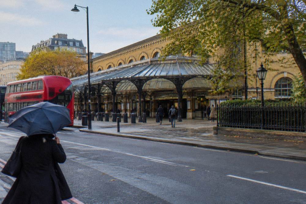 A London station exterior with a glass and brick facade and buses passing on the street