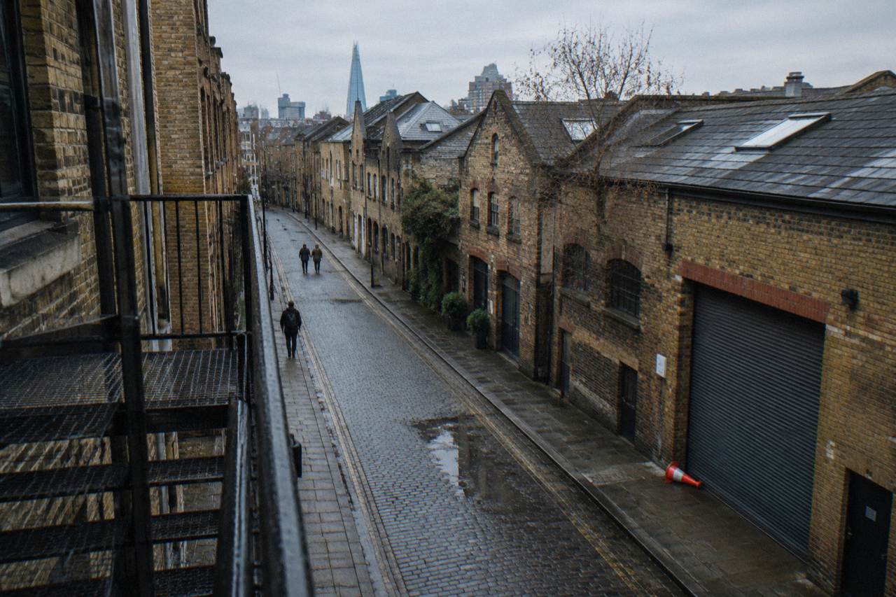 A brick-lined East London street with warehouse buildings under an overcast sky