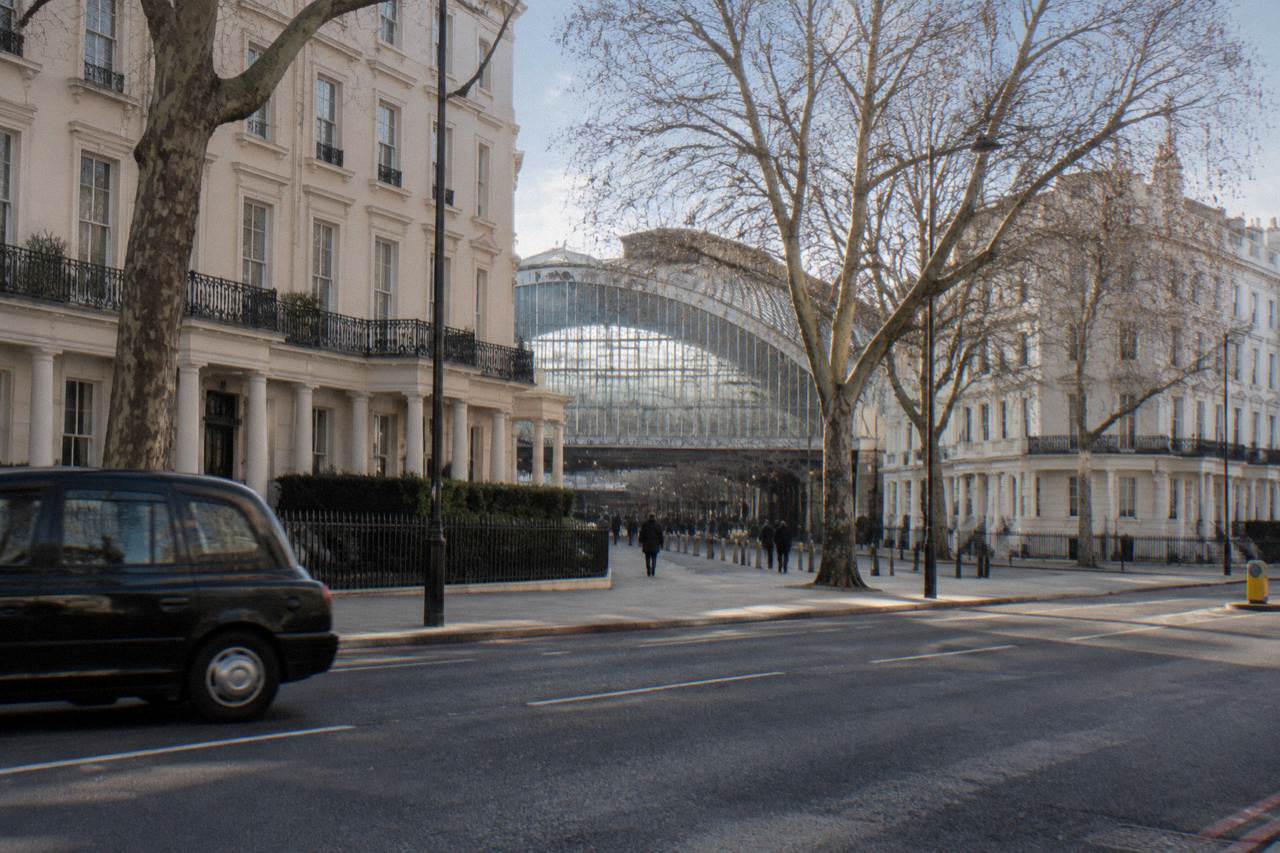 A wide London street with terraced white stucco facades and a distant station canopy roof