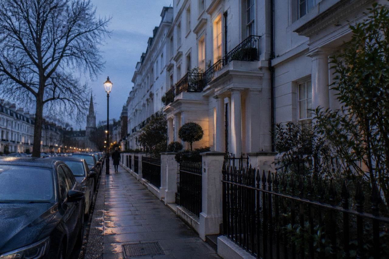 A London street of tall stucco townhouses at dusk with parked cars along the kerb