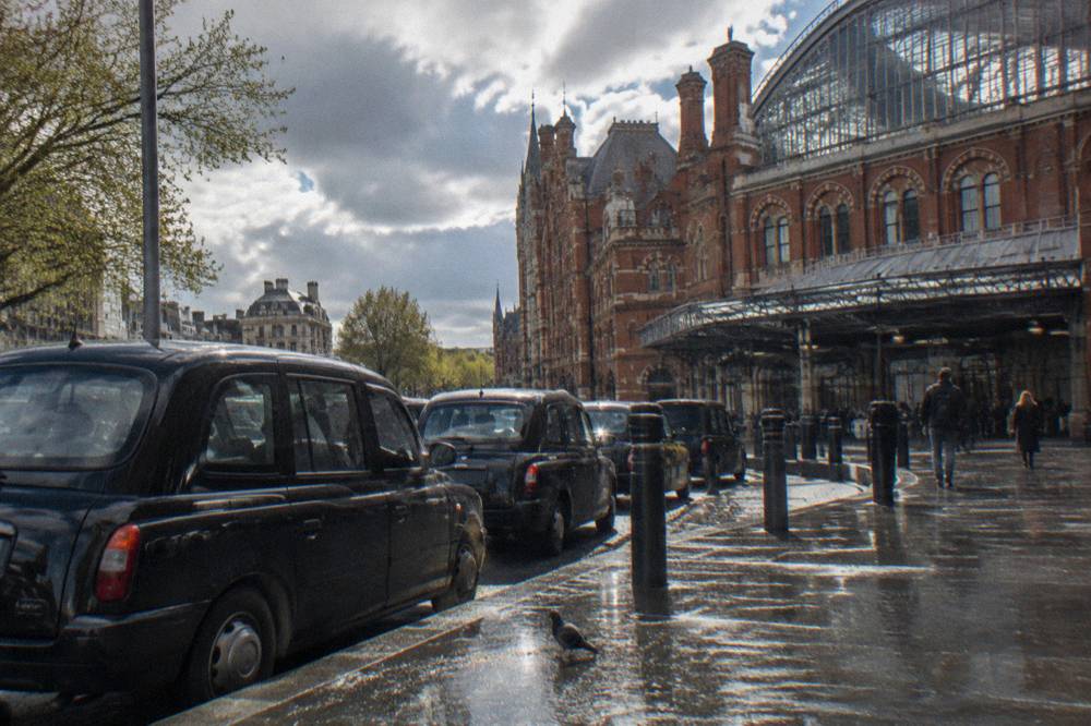 A red brick railway station exterior with a glass roof and taxis waiting outside
