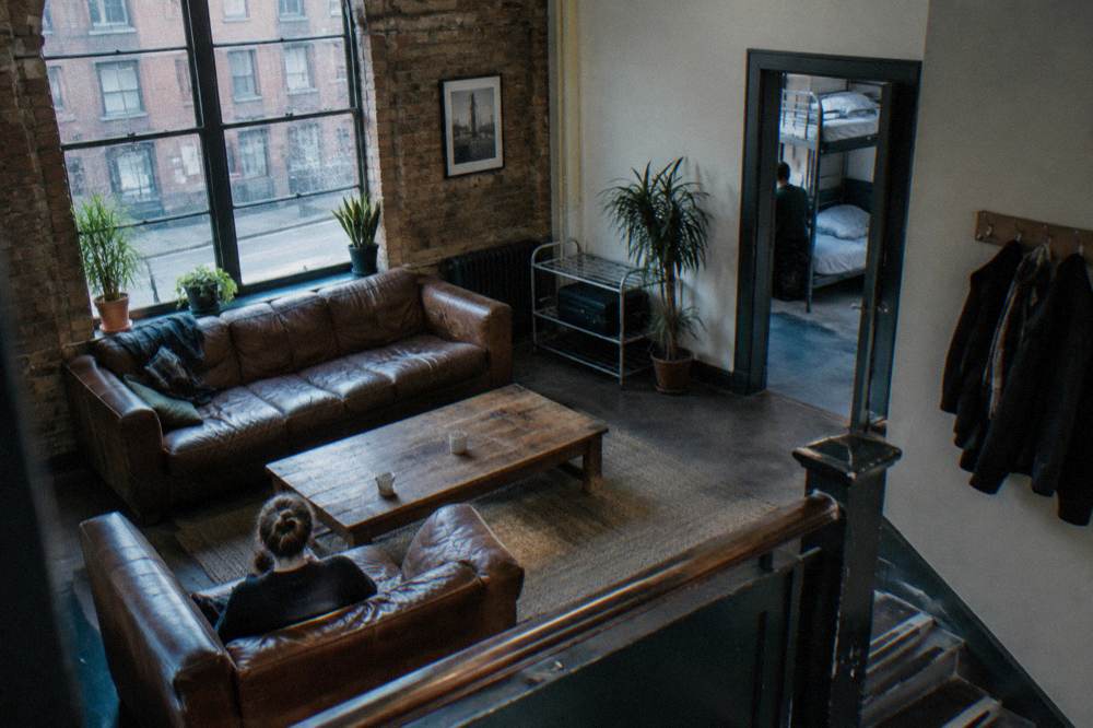 A hostel common room with sofas, a luggage rack and bunk beds visible through an open doorway